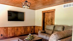 A living room with a wooden ceiling, a mounted flat-screen TV, a brown leather sofa, and a coffee table with a decorative centerpiece. - Quiet, country home with luxury amenities. (Dundee)