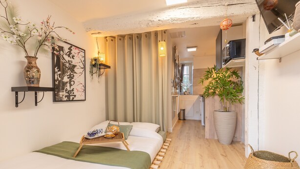 Een moderne hotelkamer met een bed, nachtkastje met dienblad, grote potplant en een wandplank met decoratieve objecten.