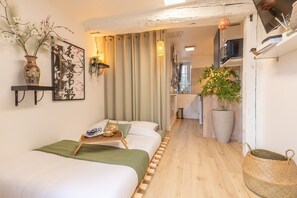 Quarto de hotel moderno com cama, mesa de cabeceira com bandeja, uma planta em vaso grande e um prateleira com objetos decorativos.