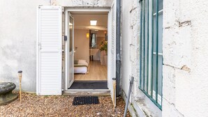 Een geopende deur die leidt naar een kamer met een bed, een klein tafeltje en een kamerplant.