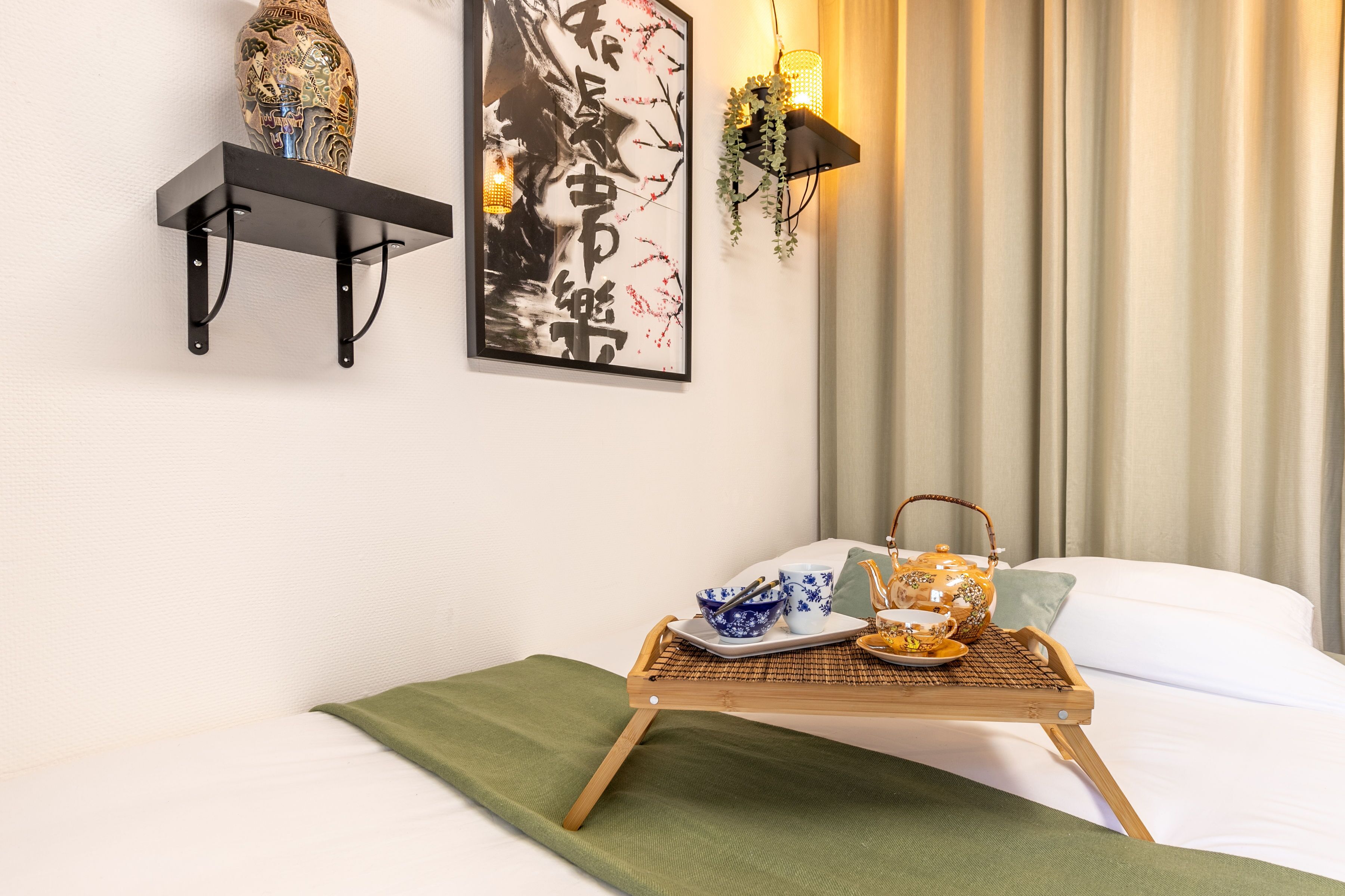 A neatly arranged hotel room with a bed, a bedside table with a tray of tea and teapot, a framed artwork, a shelf with a decorative vase, and a wall-mounted lamp.
