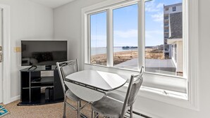 A room with a TV, a small dining table, and chairs. There is a large window with a view of the beach and buildings. - One bedroom, direct ocean views! (Old Orchard Beach)
