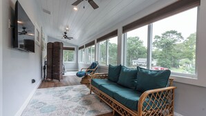 A spacious room with a large window, a ceiling fan, and a wicker sofa with teal cushions. - Seaside Serenity - New Construction! (Yarmouth)