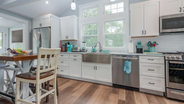 A modern kitchen with white cabinets, stainless steel appliances, and a wooden dining table. - Seaside Serenity - New Construction! (Yarmouth)
