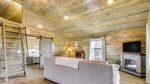 A cozy log cabin interior with a sofa, a fireplace, a TV, and a ladder leading to a loft. - Cozy Mississippi Cabin w/ Covered Porch & Grill! (Glen)