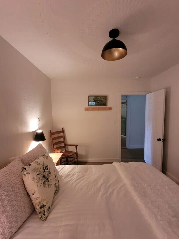 Un dormitorio con cama, una silla, una lámpara, una luz en el techo, una puerta y un cuadro pequeño en la pared.