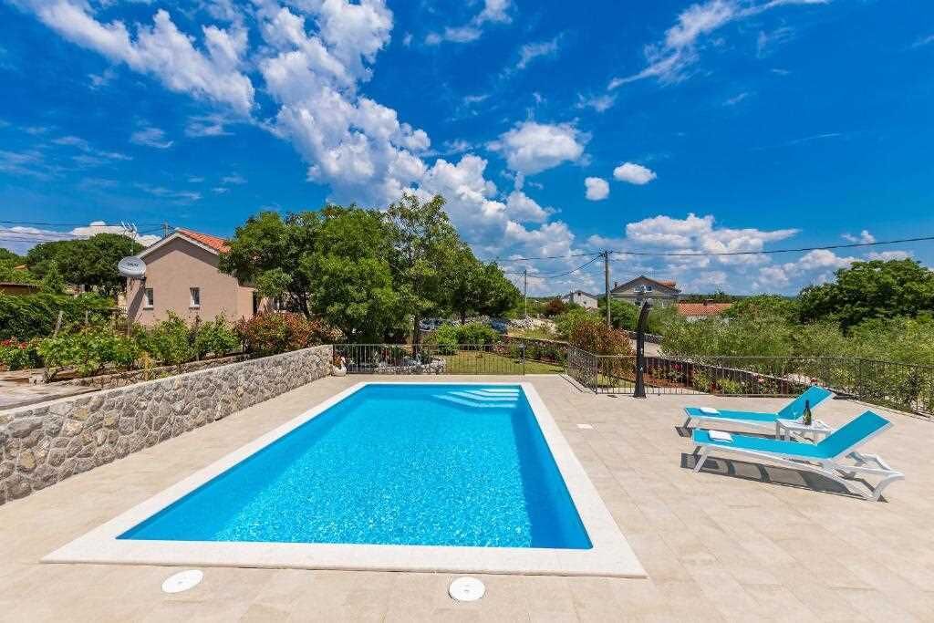 A rectangular swimming pool with a stone wall and lounge chairs, surrounded by greenery and houses.
