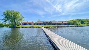 Un hotel junto al lago con un muelle de madera que se adentra en el agua, rodeado de exuberante vegetación y un cielo azul despejado.