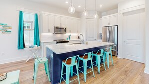 A modern kitchen with white cabinets, a large island with bar stools, stainless steel appliances, and teal accents.
