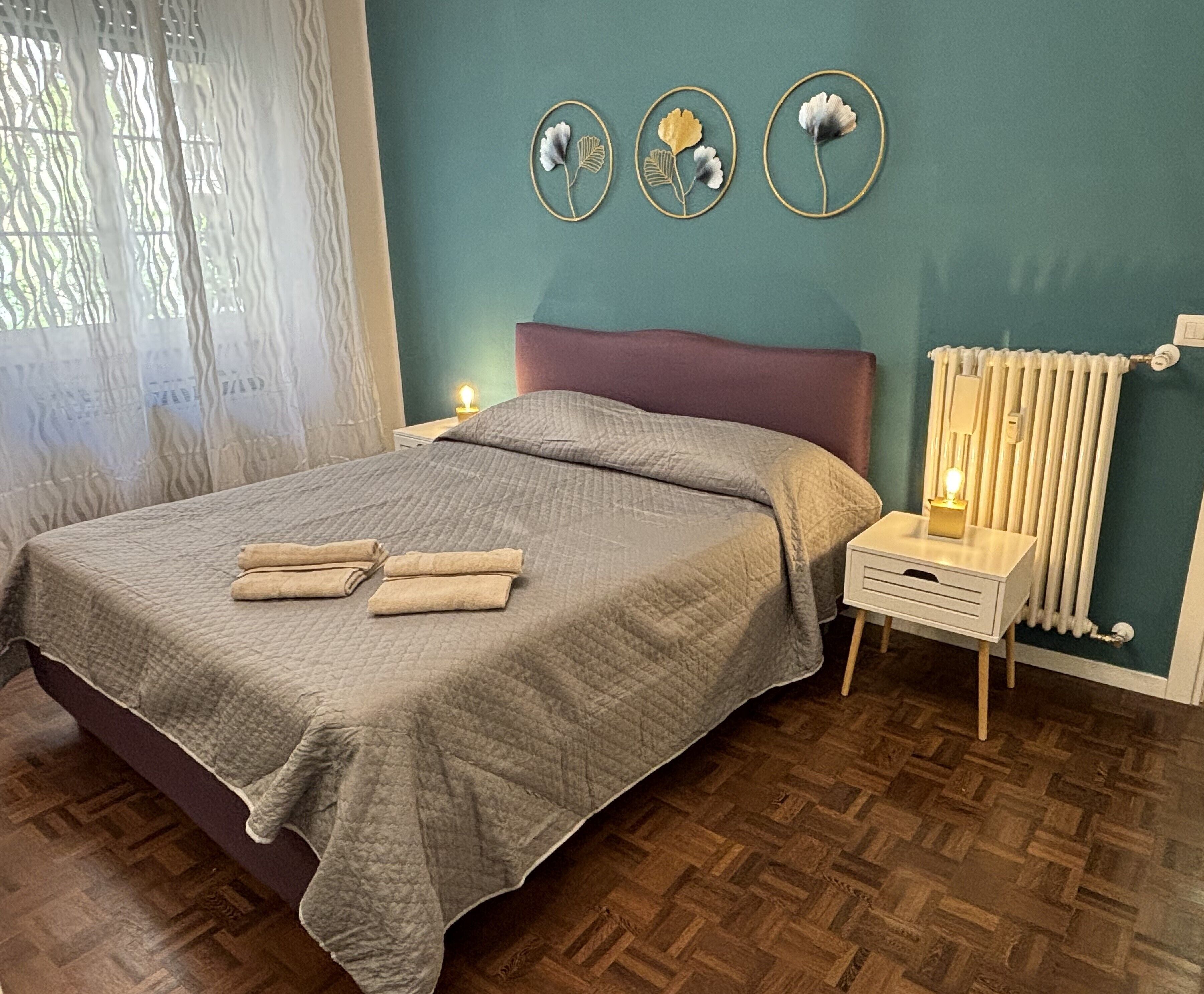 Een net opgemaakt bed met een grijze deken, een nachtkastje met een lamp en een radiator.