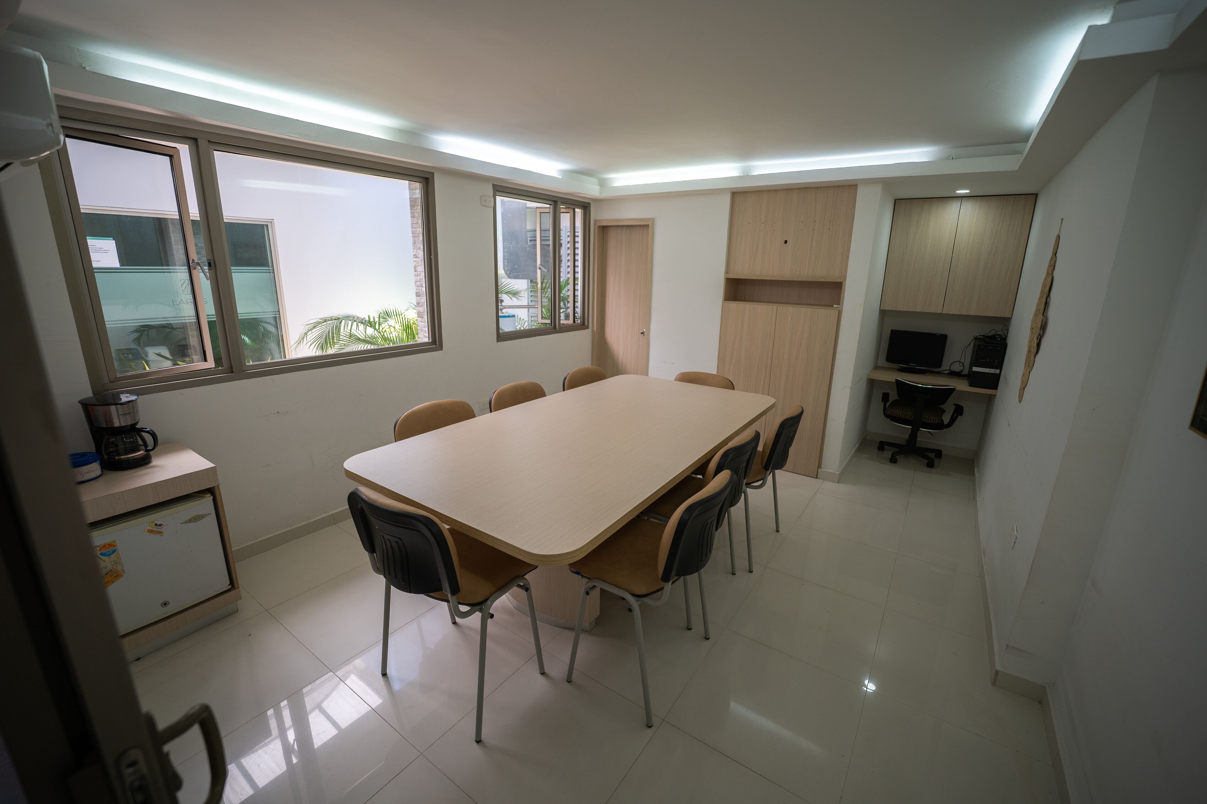 Uma sala de conferência com uma mesa comprida e cadeiras, uma kitchenette com frigorífico e máquina de café, e uma janela com vista para vegetação.