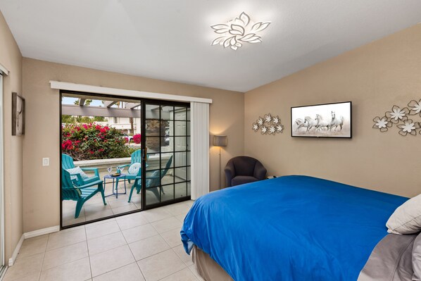 A bedroom with a large bed, a sliding glass door leading to an outdoor seating area with chairs and a table, and a TV mounted on the wall.