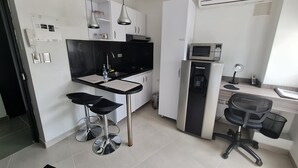 Une cuisine compacte équipée d’un four à micro-ondes, d’un évier et d’un réfrigérateur. On y trouve deux tabourets de bar et un petit espace bureau avec une chaise et un bureau.