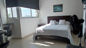 Une chambre d’hôtel équipée d’un lit, d’un bureau, d’une chaise, d’un ventilateur et d’une fenêtre donnant sur des immeubles.