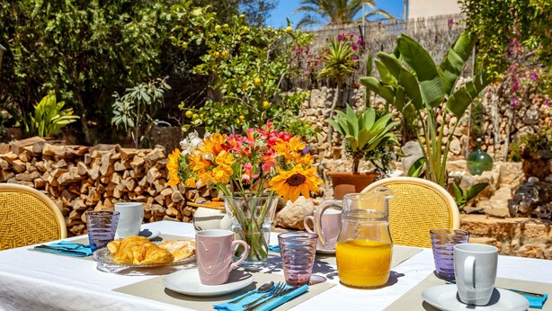 An outdoor dining table set with a croissant, coffee cups, a pitcher of orange juice, and a vase of flowers. - Can Juanito by dracmallorca (Cala Llombards)