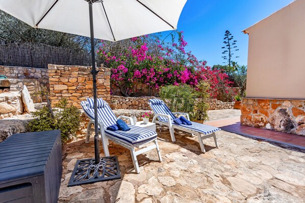 Eine Terrasse mit zwei gestreiften Liegestühlen, einem großen Sonnenschirm und einer Steinmauer, die mit rosa Bougainvillea bepflanzt ist.