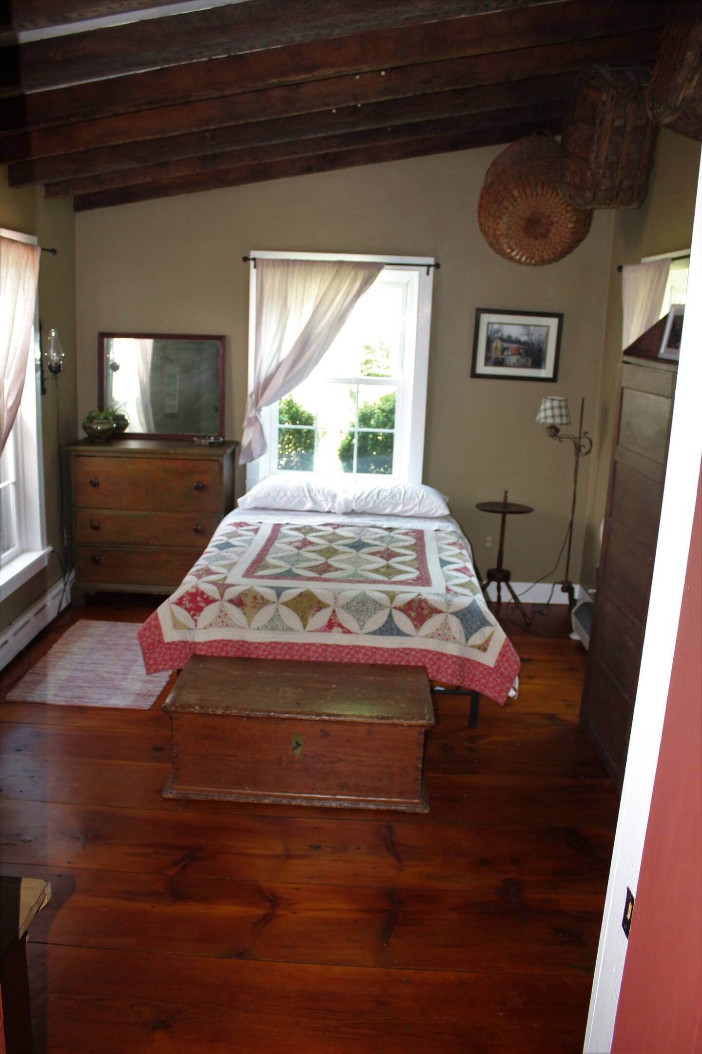 A bedroom with a wooden floor, a bed with a quilt, a dresser, a window with curtains, and a lamp.