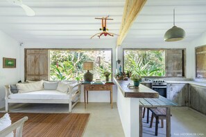 A rustic kitchen with wooden cabinets, a dining table, and a sofa. There are windows with views of greenery outside. - Casas da Lia (Porto Seguro)