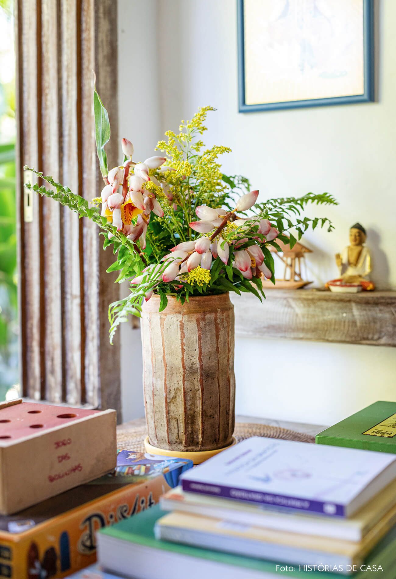 A rustic wooden vase with flowers on a table, surrounded by books and a Buddha statue.