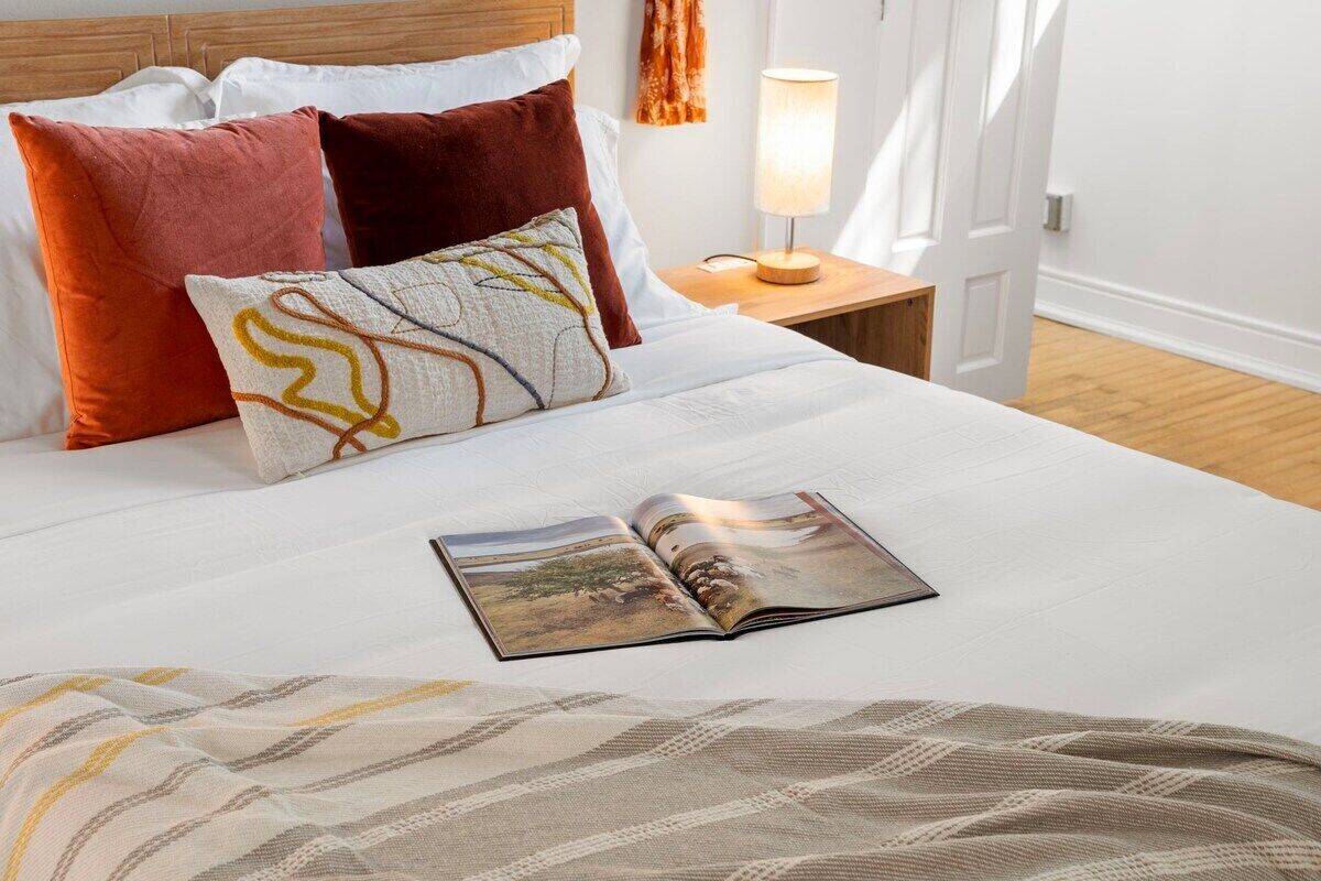 Cama bem arrumada com revista, almofadas e abajur em uma mesinha de cabeceira.