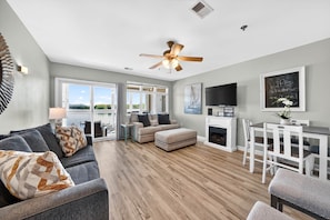 A spacious living room with a grey sofa, a ceiling fan, a fireplace, and a dining area with white furniture. - #233 Pointe - 2 Bedroom Lakefront Condo at Mariners SML (Bedford County)