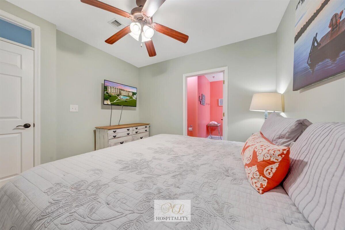 A bedroom with a bed, a ceiling fan, a television, a dresser, and a door leading to another room.