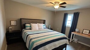 A bedroom with a bed, a nightstand, a bedside table, a ceiling fan, a window with curtains, and a wall with a framed picture.