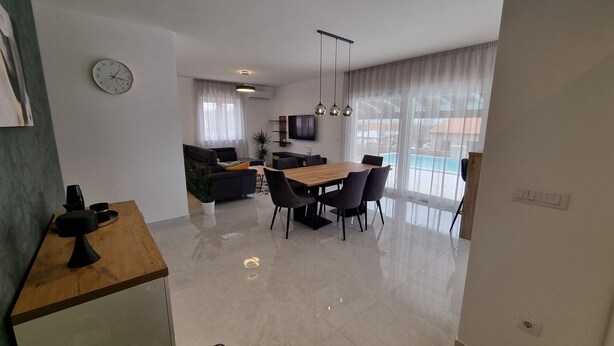 A modern living room with a sofa, a dining table with chairs, a TV mounted on the wall, and a large window with sheer curtains. - Beautiful newly built villa with private pool in a small village in Istria (Sveti Petar u Šumi)