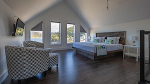 A spacious bedroom with a large bed, a seating area with patterned chairs, and multiple windows offering views of the outdoors. - The Tides Inn (Norris Point)
