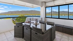 Un balcon avec une table et des chaises pour prendre les repas, un ventilateur de plafond et une vue sur un lac, avec des bateaux et des montagnes en toile de fond.