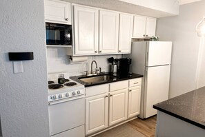 Una cocina con armarios blancos, encimera negra, pileta de acero inoxidable, estufa y refrigerador.