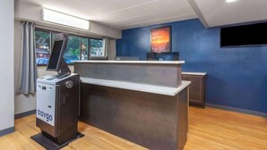Un mostrador de recepción con un quiosco digital, una pared azul con una pintura de atardecer y una ventana con cortinas.