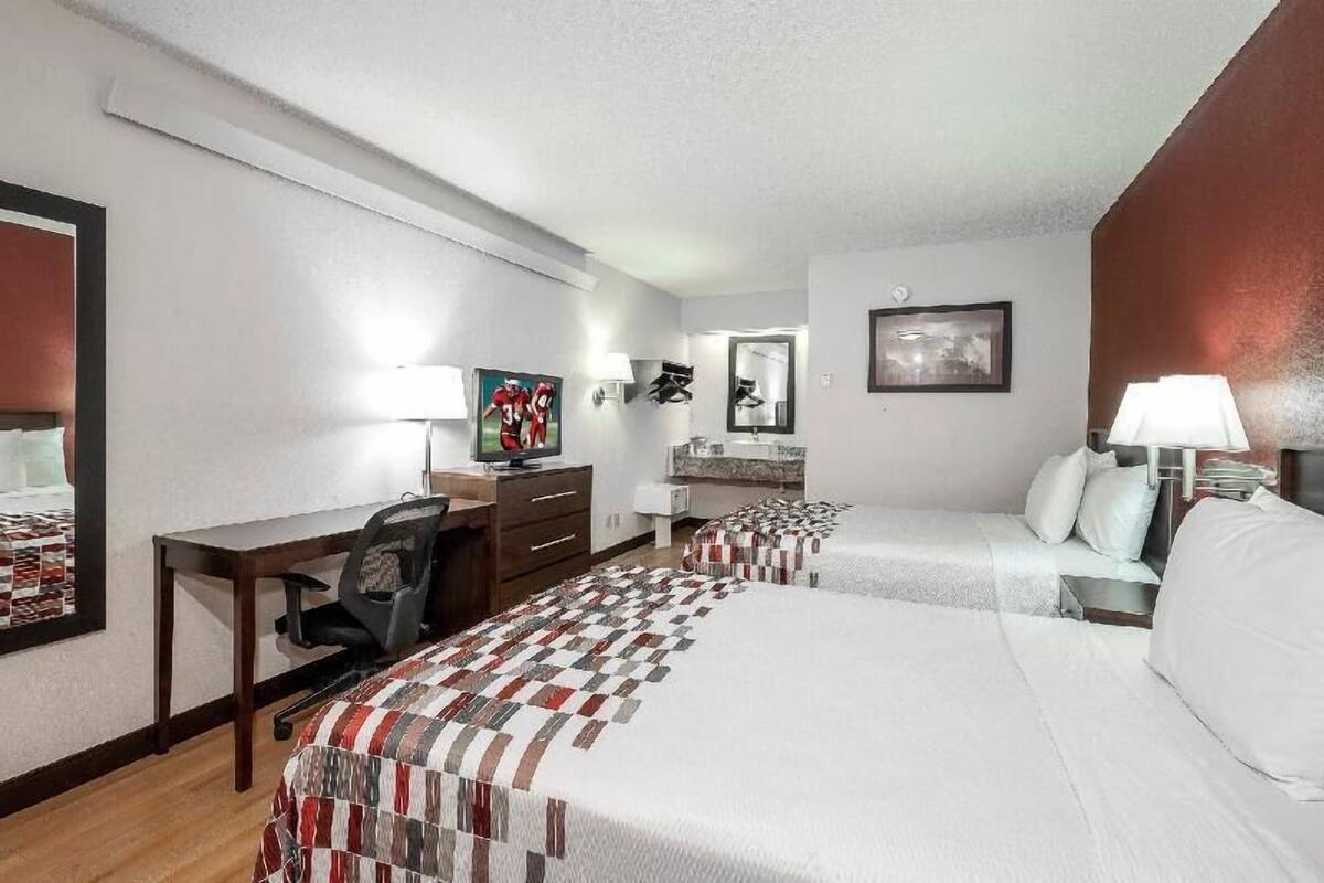 A hotel room with two beds, a desk, a chair, a TV, and a bathroom mirror.