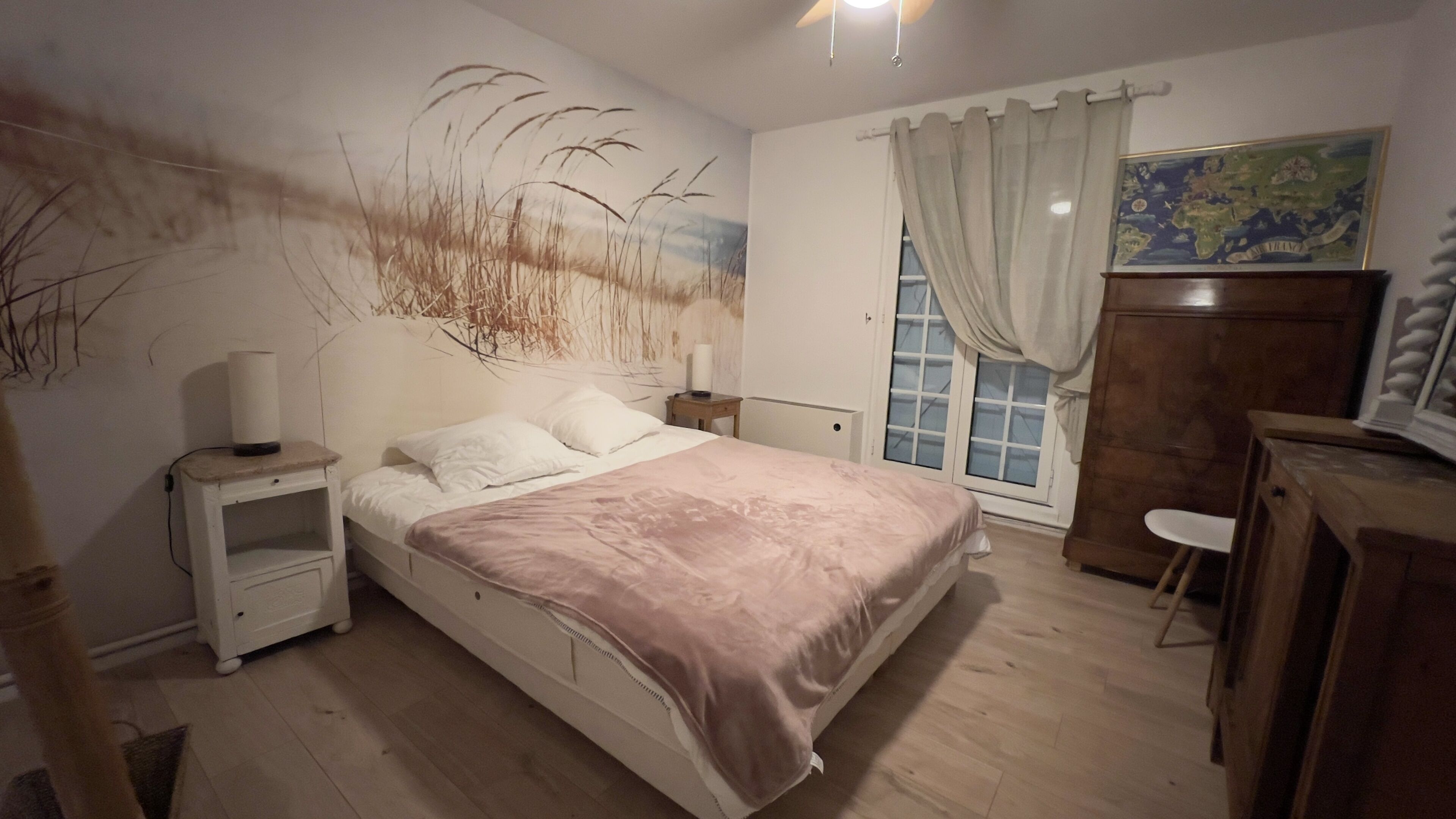 A bedroom with a bed, bedside tables, a dresser, a window with curtains, and a wall mural.