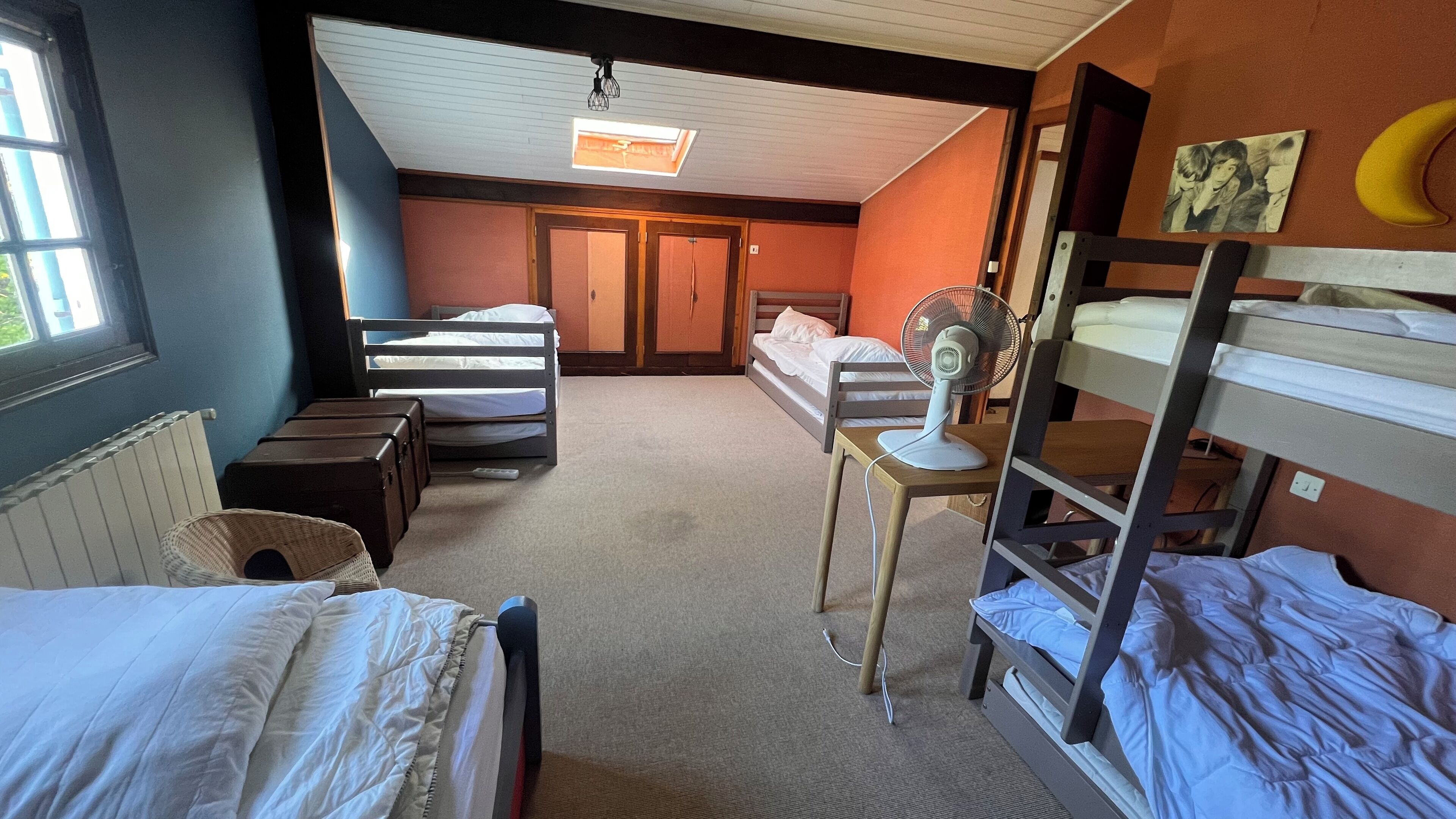 A dormitory room with bunk beds, a desk with a chair, a fan, and a window.