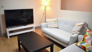 A living room with a flat-screen TV on a white stand, a white sofa with striped pillows, a black coffee table, and a floor lamp. - Pleasant Apartment in City Center (Birmingham)