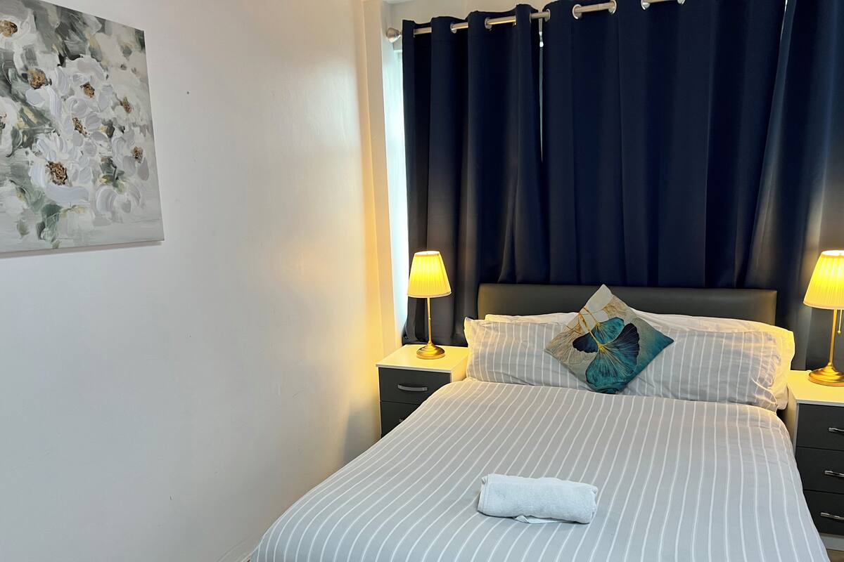 A neatly made bed with striped bedding, a folded towel, and a decorative pillow. There are two bedside lamps on nightstands, and a dark curtain covers the window.