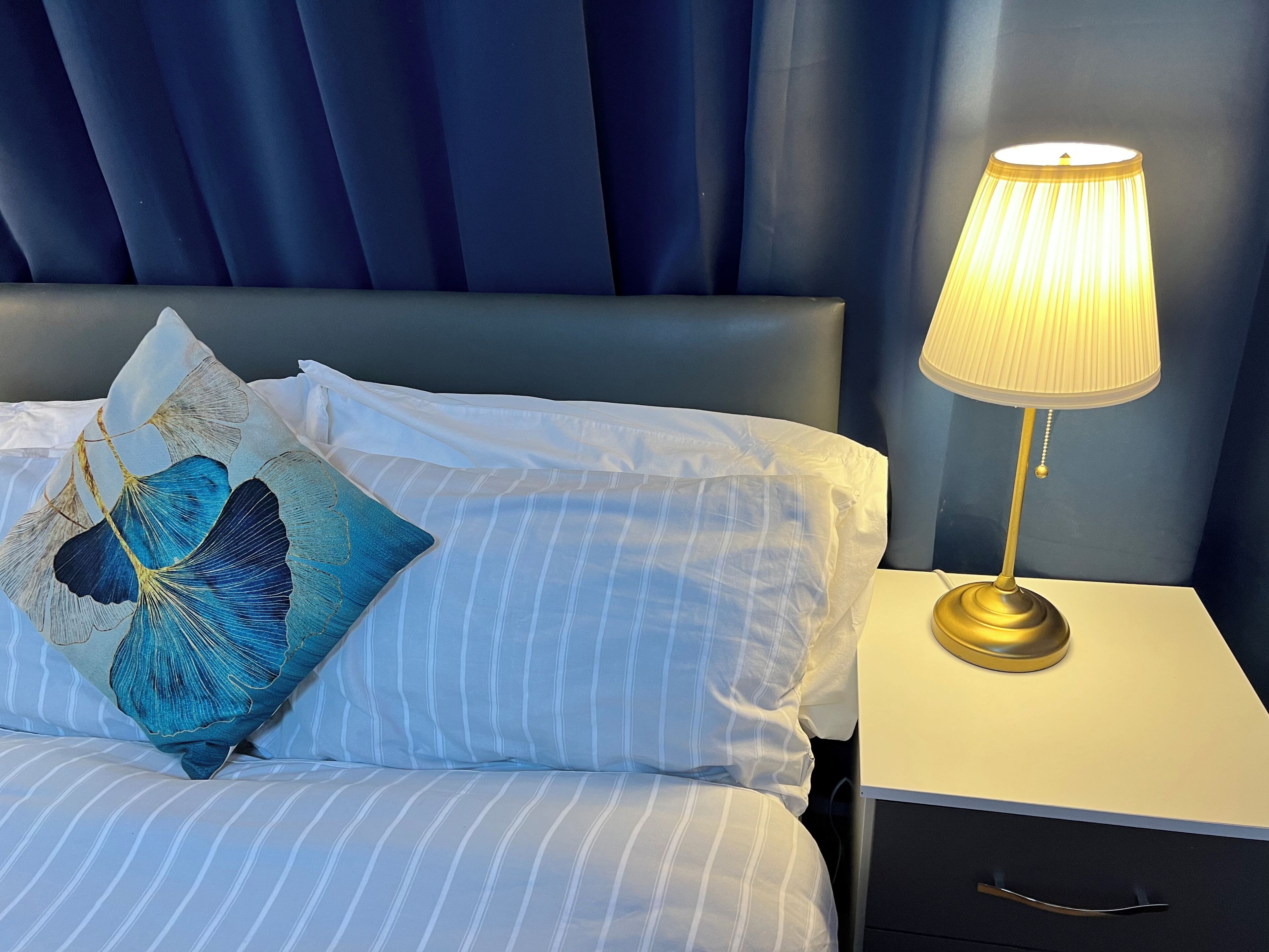A bed with striped bedding and decorative pillows, a bedside table with a lamp, and a dark blue headboard.