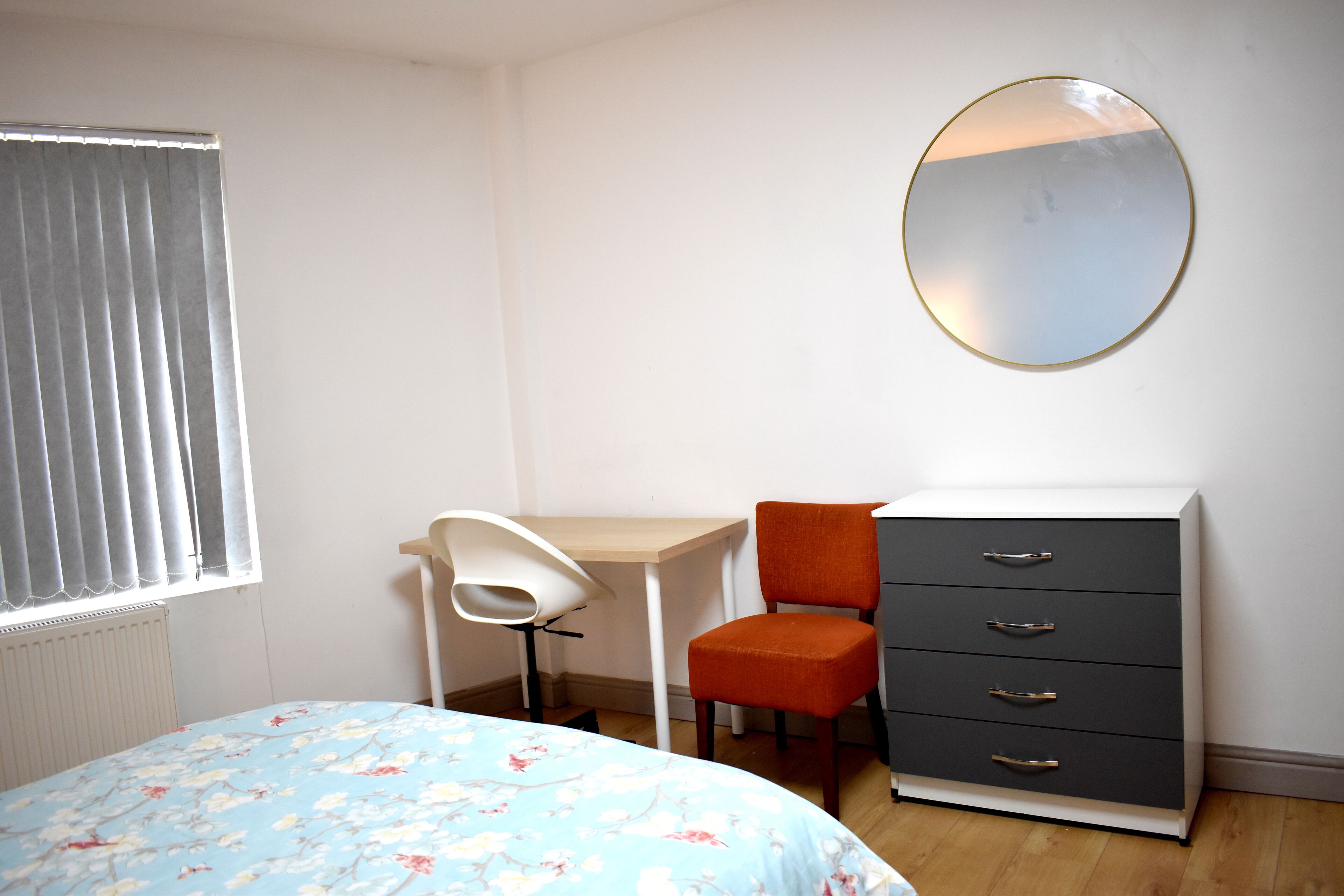 A bedroom with a bed, a desk, an orange chair, a dresser, and a round mirror on the wall.