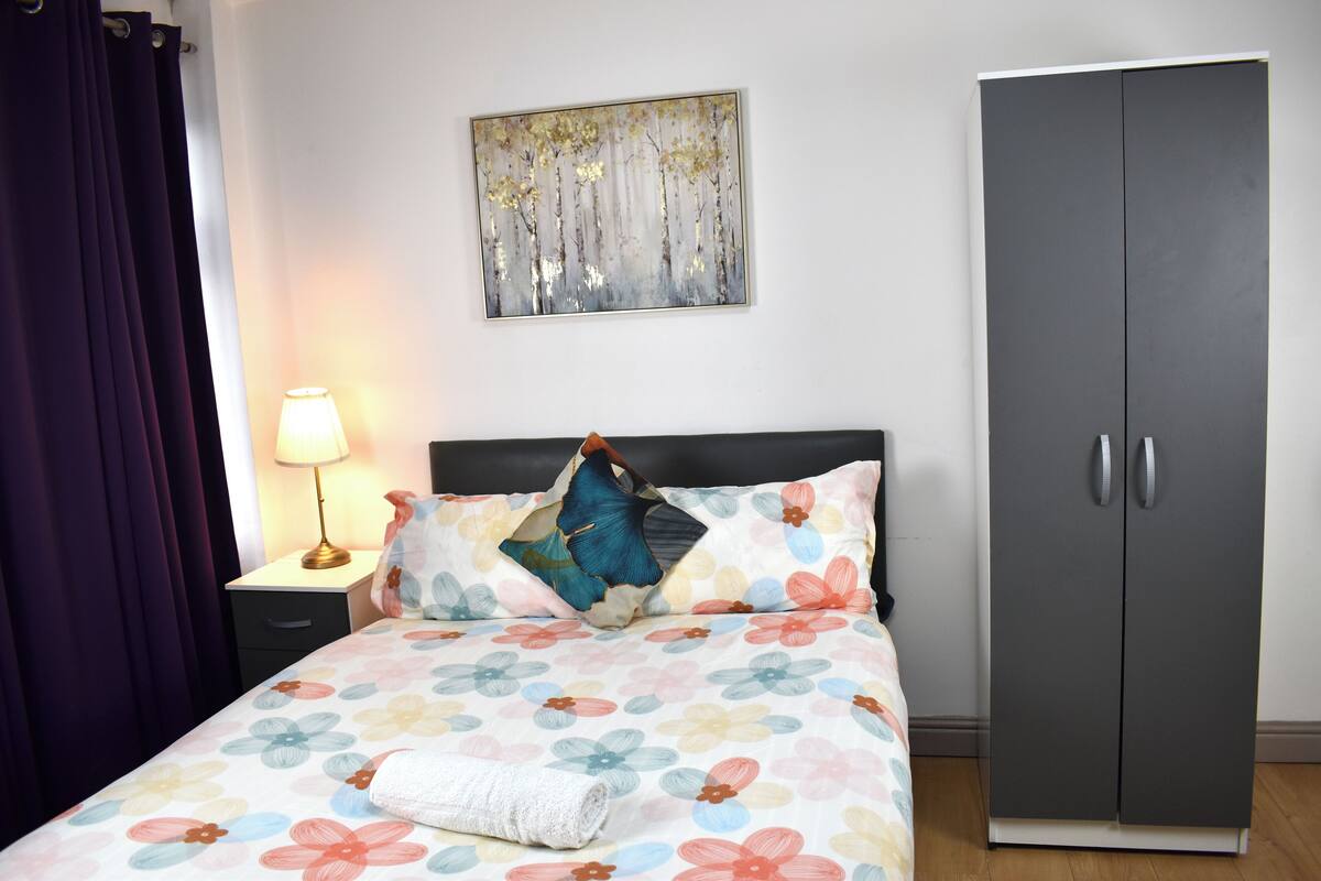 A bedroom with a bed, a nightstand, a lamp, a wardrobe, and a painting on the wall.