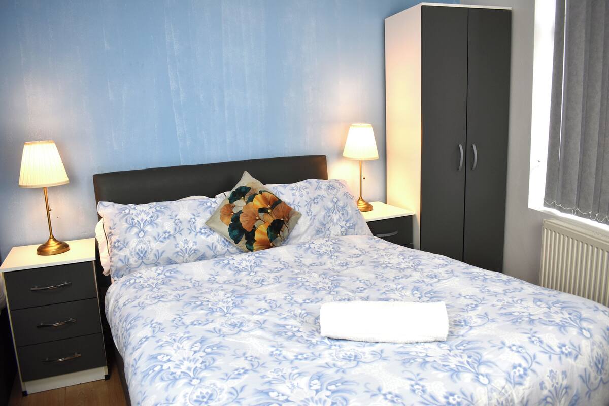 A neatly made bed with a floral bedspread, two bedside lamps, a dark headboard, and a wardrobe.