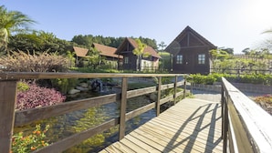 A wooden deck with a small pond and a bridge, surrounded by tropical plants and a building with a red roof. - Pousada Scursel (Ibirama)