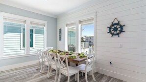 A dining area with a wooden table and white chairs, large windows with blinds, and a wall-mounted ship's wheel decoration. - Here and Now | 🛥️ Canal Access + Pool + Beach Gear + Golf Cart + Sleeps 20 (Navarre)