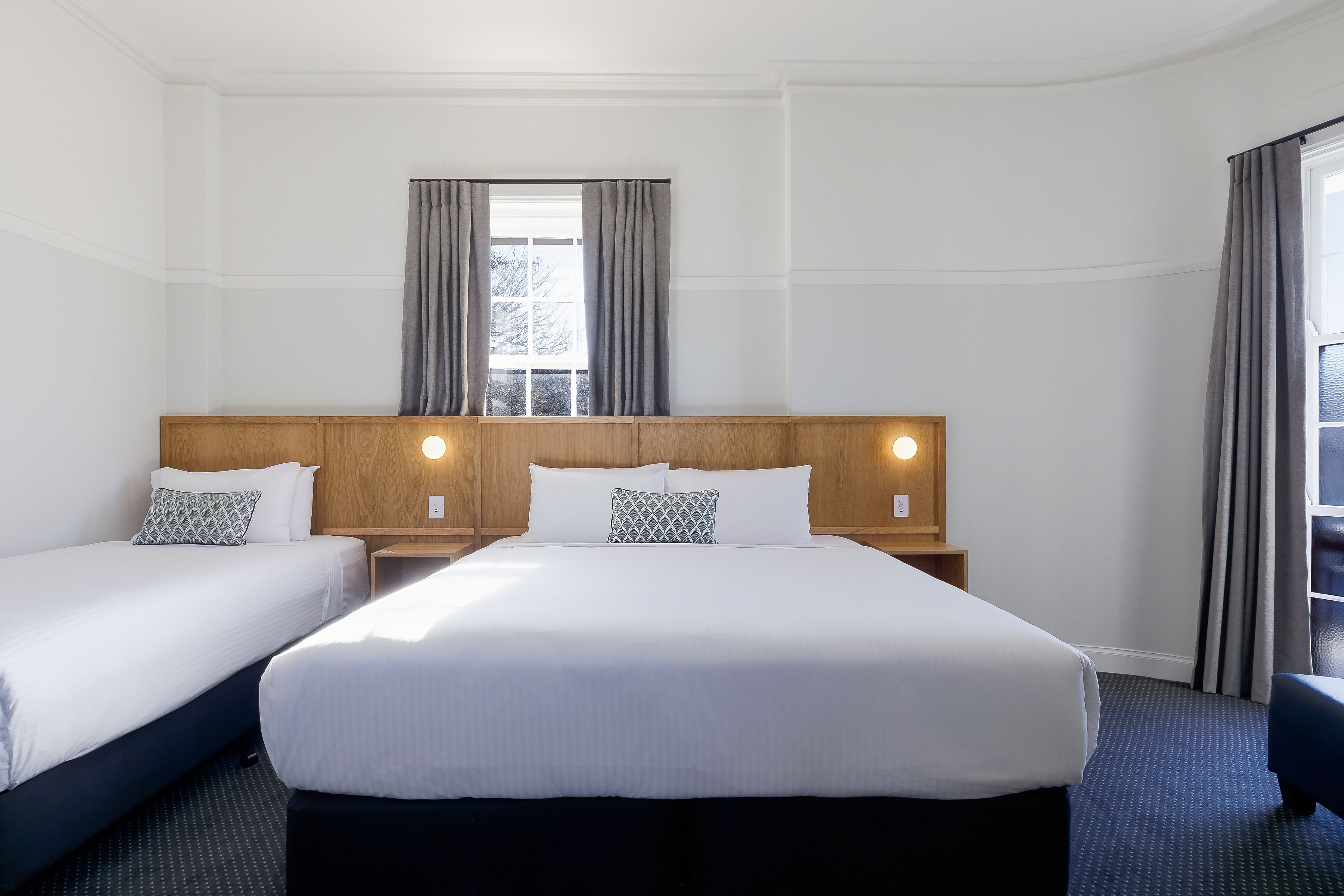 Une chambre d’hôtel avec deux lits, une tête de lit en bois, une fenêtre avec des rideaux et une table de chevet avec une lampe.
