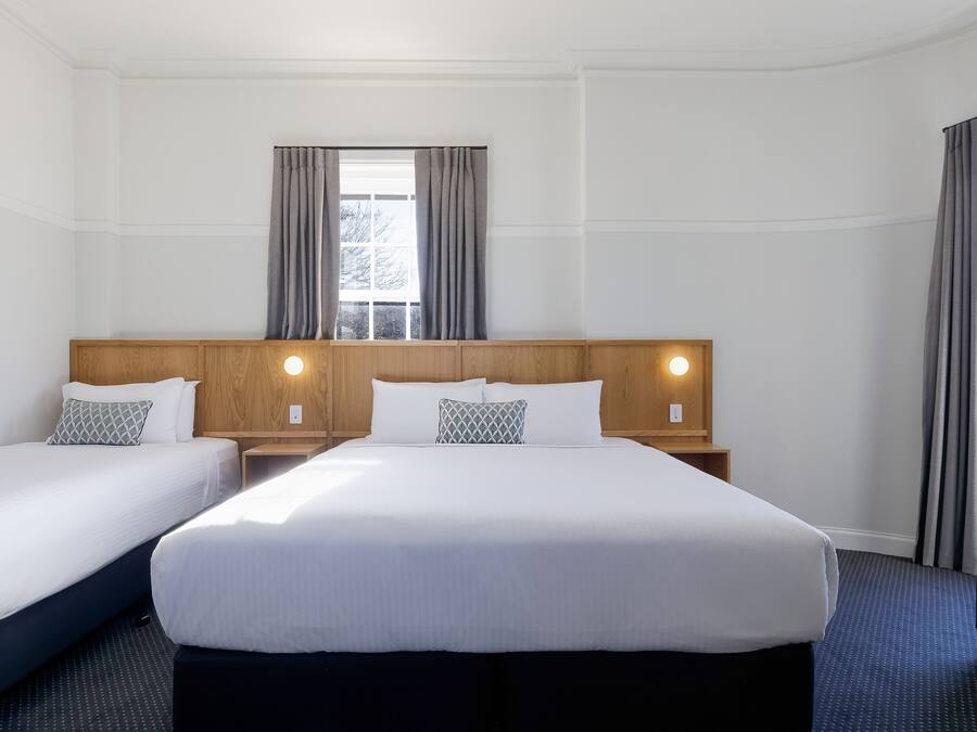 A hotel room with two beds, a wooden headboard, a window with curtains, and a nightstand with a lamp.