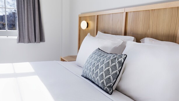 A neatly made bed with white linens and a patterned pillow, a wooden headboard with a light fixture, and a window with curtains. - Mortdale Hotel (Mortdale)