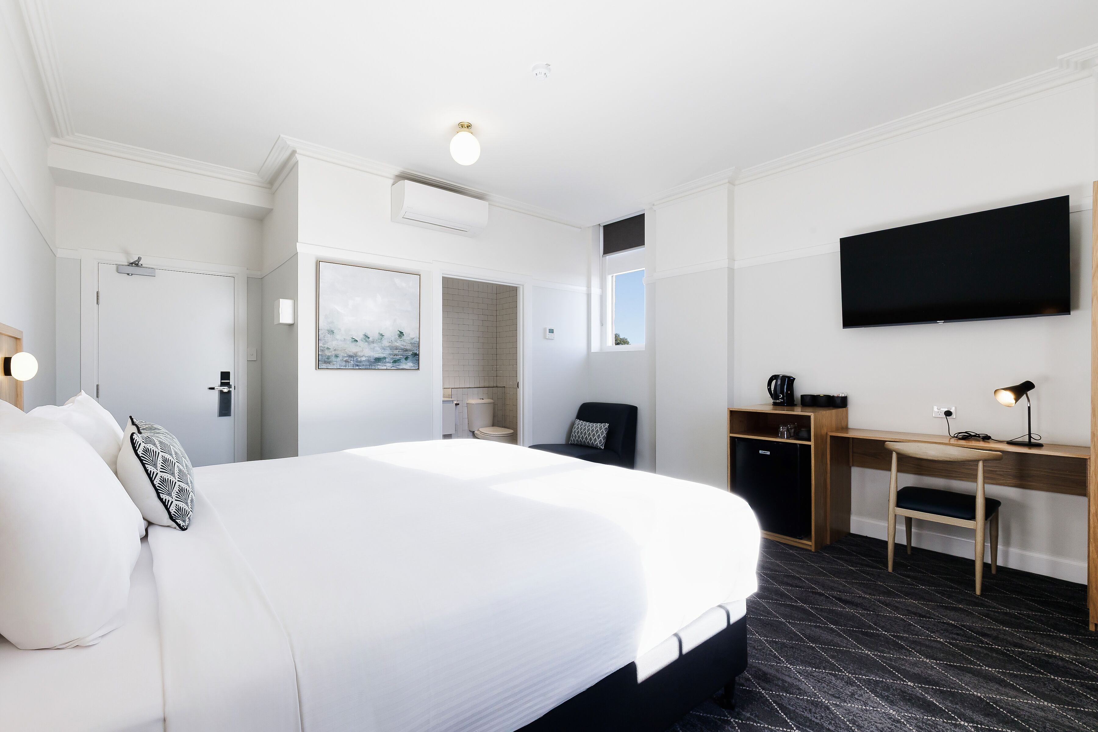A hotel room with a large bed, a desk, a chair, a flat-screen TV, and a coffee machine.