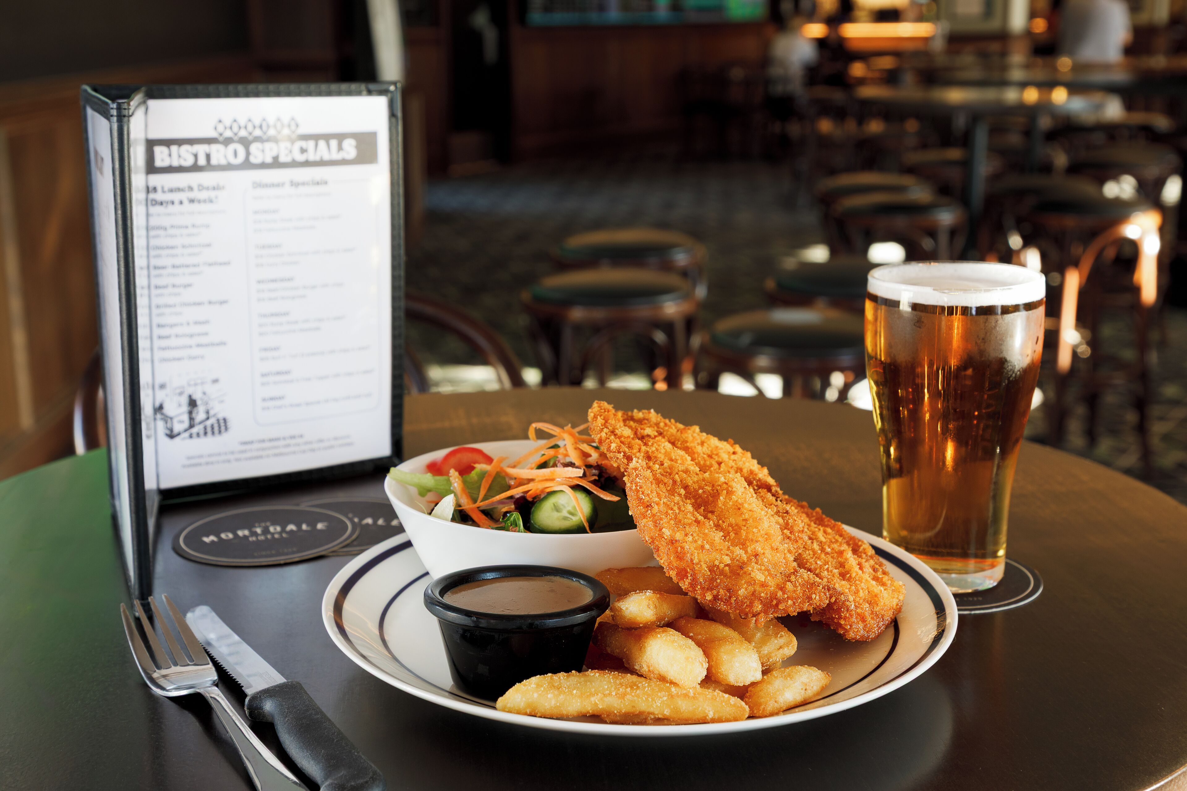 Un panneau affichant le menu, intitulé « SPÉCIALITÉS DU BISTROT », présente une assiette de nourriture comprenant du poisson pané, des frites et une salade, ainsi qu’un verre de bière et une petite coupelle de sauce.