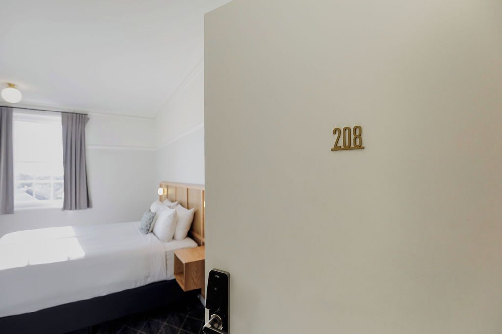 Habitación de hotel con cama, mesita de noche, ventana con cortinas y el número 208 en la pared.