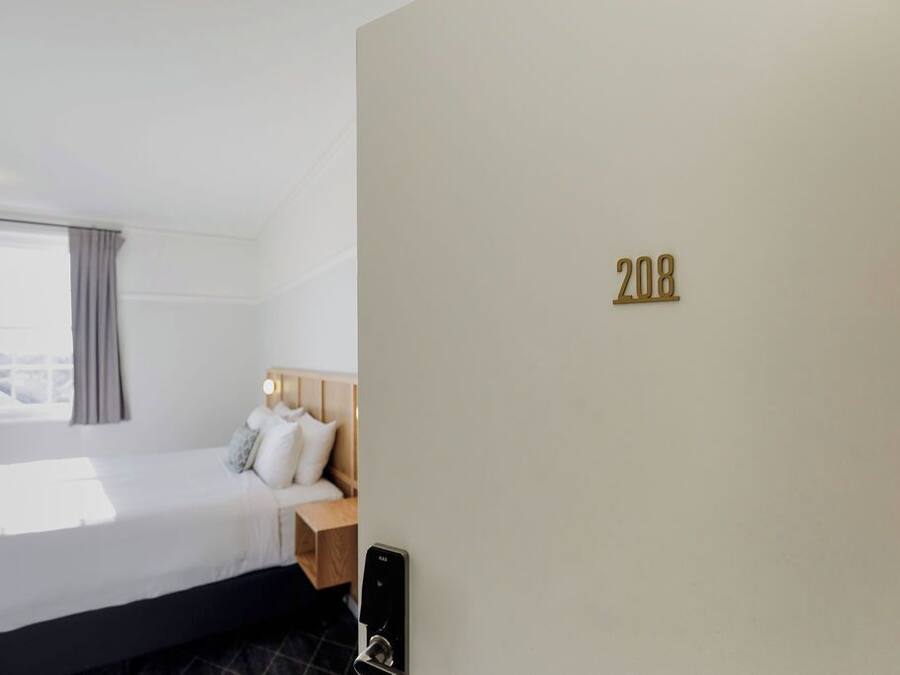 Habitación de hotel con cama, mesita de noche, ventana con cortinas y el número 208 en la pared.
